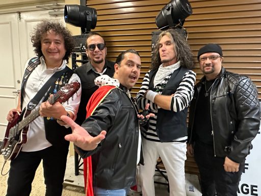 Five musicians in stylish outfits posing together, two with guitars and unique hairstyles.